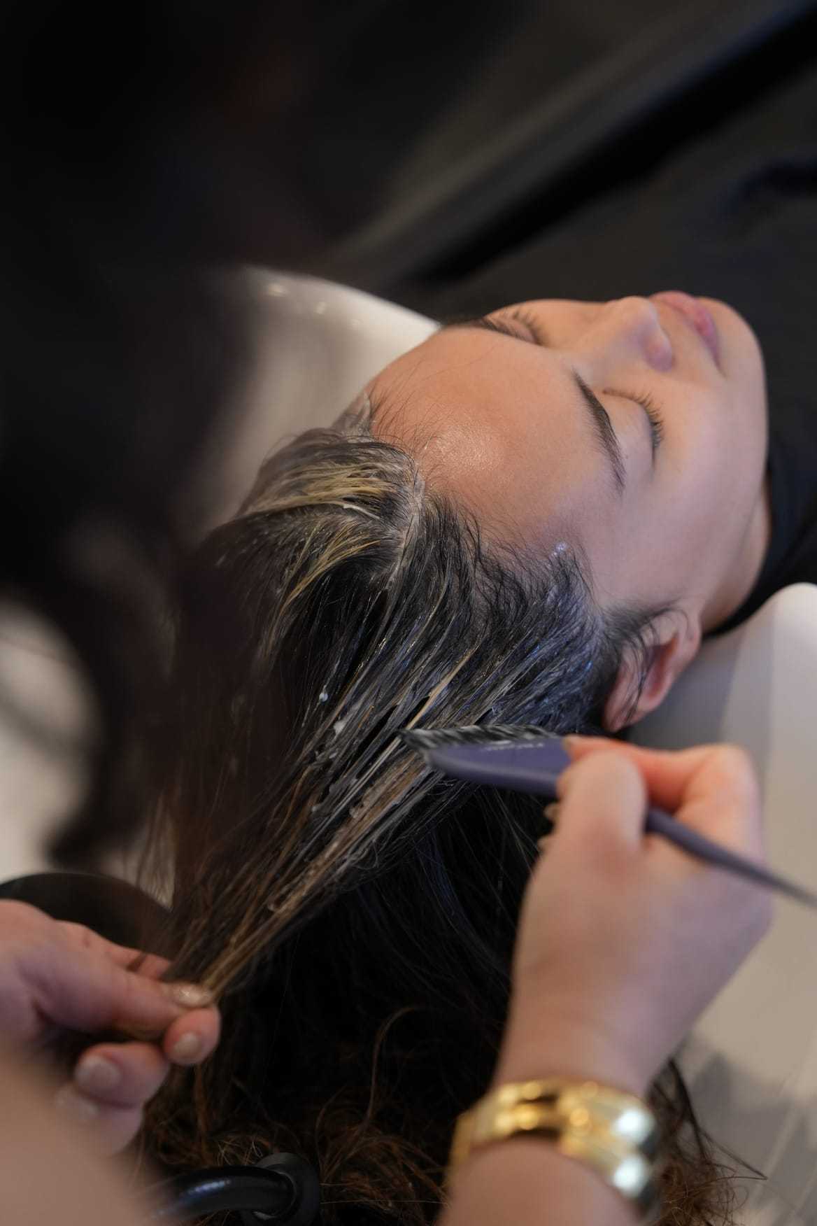 Hair treatment application at a salon, person reclining with stylist applying product.